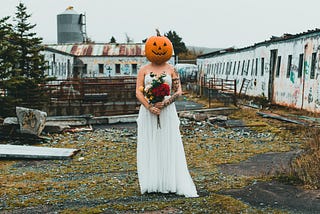 a woman with a pumpkin head wearing a white off-shoulder dress and holding a red and yellow flower bouquest is standing in a discarded place with rows of white boarded up house behind her