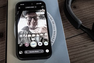 Photo of an iPhone open to the TikTook app, resting on a book. The app has pulled up a photo of a man wearing a hat and glasses who is making a cutting motion at his neck.