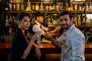 An attractive man and woman are sitting at a bar, looking directly at the viewer, seductively raising their cocktail glasses as if inviting the viewer to join them.
