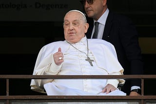 Pope Francis, seated in a wheelchair, gives a thumbs-up to the crowd from the balcony of Gemelli Hospital in Rome.