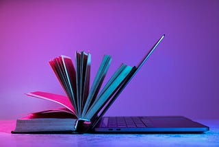 Livro aberto encostado em um laptop semiaberto, iluminados por luzes coloridas em tons de rosa e azul, sobre uma superfície plana.