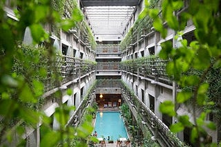 We’re looking at the inside of a large building. Skylights flood the space with natural light. The walls are covered in fresh green vines and leaves. There’s a swimming pool in the middle of the space. There are loungers at the far end of the pool and tables and chairs at the end nearest to us. People are sitting at the tables.