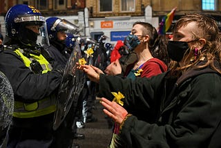 Police in riot gear with helmets and plastic shields in confrontation with those on the left of the image: young people wearing face masks. They are outdoors in a street, shops faintly seen in the background.