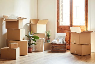 Cardboard moving boxes stacked in a sunlit room with plants and a wooden door.