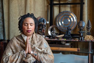 photos Aishah, brown skinned Black woman, wrapped in a golden shawl, with palms together, and eyes closed. She is seated in front of an altar with statues of West African Deities, Buddha, Quan Yin, and a gong.