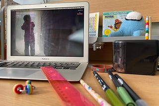 A laptop on a desk with a photo of a child in a storm, yelling on the desktop. There are pens and pencils on the desk, a stim toy and a ruler. In the back is a French book about winter and a stuffed Among Us video game character.