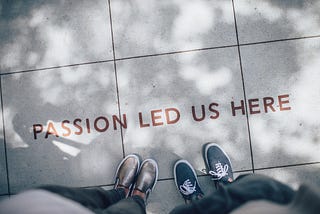 A view of 2 sets of feet, seen from above, standing on a sidewalk. The words “Passion Led Us Here” are printed on the sidewalk.