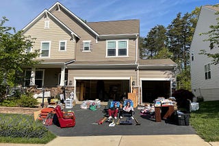 Photo of a beige two-story home with three car garage, doors open. Yard sale item in the driveway. In the center, two grown children smiling.