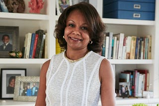 Woman posing with a smile in white dress in front of bookshelves with books and family photos.