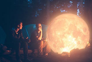 Two young man sitting next to a glowing campfire that resembles a yellow moon.