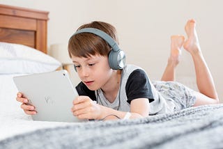 White male child laying on a bed on his stomach with his feet in the air holding a tablet with headphones on.