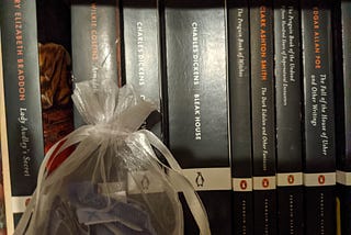 Eight Penguin Classics paperbacks — the ones with black spines with titles in white and author names in white or orange-red. To the left, there is a small lacy bag with a ribbon that contains tiny pieces of paper.