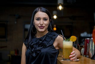 Image of girl in black blouse and hoop earrings holding a cocktail at a bar