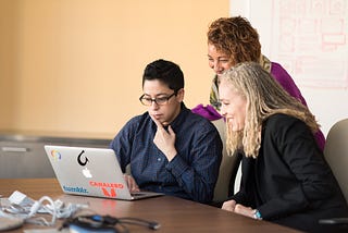 three employees collaborate around a laptop computer sitting on a table