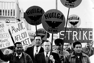 Black and white photo of Larry Itliong, Cesar Chavez and others holding signs that say, “Huelga NFWA,” “strike,” and “AWOC AFL-CIO Picket” during the Delano Grape Strike of 1965