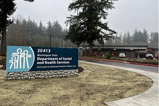 A photo of the residential treatment center at Maple Lane, with a sign that is branded with the Washington State Department of Social and Health Services, its logo and a new one-story building with trees in the background.