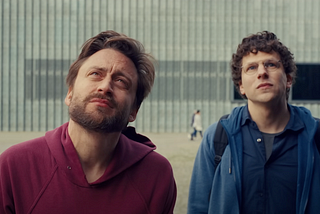 Jesse Eisenberg and Kieran Culkin look upward with serious expressions in front of the POLIN Museum’s glass-panel façade in A Real Pain (2024).