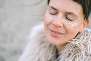 A young woman with glitter on her cheeks smiles gently with closed eyes