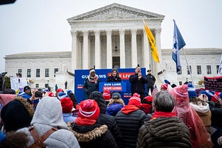 A crowd gathers in front of the Supreme Court for a rally. Signs and banners say, “gun laws save lives”