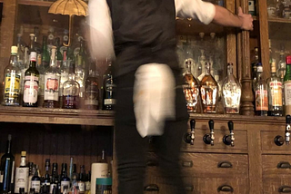 A bartender standing on a back bar about to pick a bottle of spirits