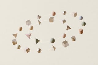 Conjunto de formas geométricas em tons pastéis — esferas, cubos, cones, cilindros e pirâmides — organizadas em dois anéis elípticos entrelaçados sobre um fundo bege claro, criando a aparência de órbitas flutuantes e harmônicas.