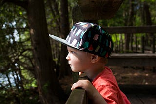Toddler looking over fence with baseball cap on towards the sunlight.