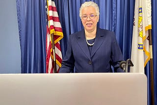 Cook County Board President Toni Preckwinkle stands behind a podium delivering a virtual keynote address to the League of Women Voters of Cook County. She is wearing her glasses, a pearl necklace, and navy blue shirt and blazer. On the sides are the American Flag and the Cook County flag. A laptop can also be seen.