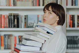 Woman wearing white with short dark hair holds a pile of books that reach her chin. She stands in front of white bookshelves lined with books and she is looking at the camera.