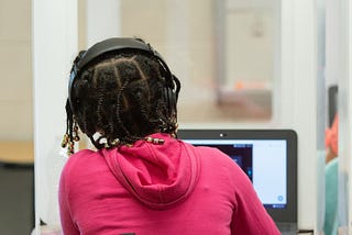 A girl in a pick sweatshirt wears headphones and types on her laptop while seated at a desk behind a protective screen