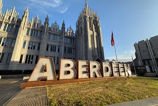 A large sign saying “Aberdeen” is written in large letters on a patch of grass. Behind it is a large building with Gothic architecture. The sky is blue.