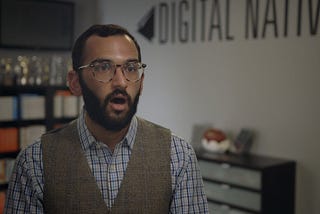 Foreground: Chest-up shot of the author, Jonathan. He is wear a blue plaid collared shirt with a gray vest over it and his mouth is ajar. Background: Several cabinets and a gray wall with the Digital Natives logo