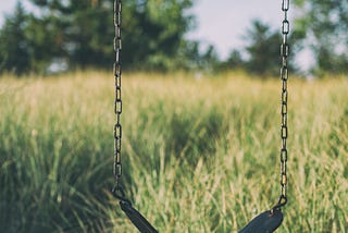 A black swing connected to a chain link hangs in a an empty field of grass
