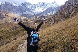 A woman with her arms raised on a trail headed towards a mountain range.
