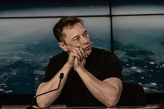 Elon Musk in black shirt with hands joined on table touching face and in front of a mike.