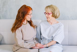 A red-haired woman smiles at a blond-haired woman as they sit on a sofa together, holding hands