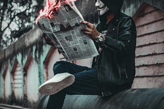 Man wearing hoodie and leather jacket with a white mask, sitting on a metal pipe, reading a newspaper that is on fire.