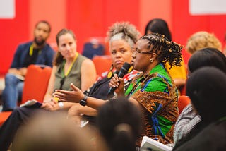An African woman holds a microphone and speaks while a small seated crowd listens