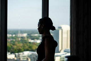 A woman is standing by a window in what appears to be an office building. She is looking out thoughtfully.