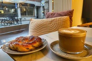 A cardamom bun and a coffee on a cafe table with a view of a Danish street behind.