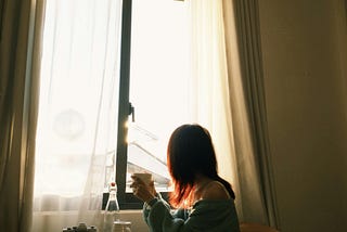 A woman sits beside a sunlit window holding a warm drink as soft morning light fills the room, conveying peace, silence, and contemplative presence.