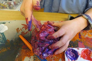 A child’s paint covered hands holding a large ball of cubed paper, which is covered in mixed colors of paint on top of a sheet of paper covered in multi-colored smears of paint.