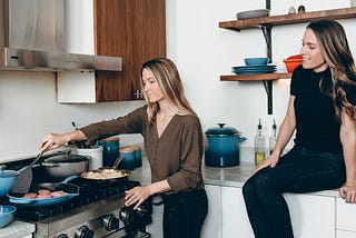 two female friends in a kitchen. One is standing at the stove, cooking. the other is sitting on the counter behind her, looking on.