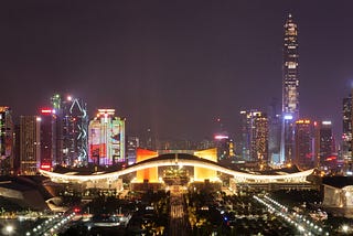 The cityscape of Shenzhen at night, with a purple black background sky and buildings with white and yellow lights.