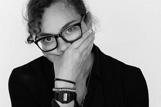 A young woman in glasses covers her mouth with one hand, eyes wide and thoughtful. She wears a black outfit and digital watch, standing against a plain background, appearing amused, uncertain, or deep in thought.