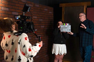Man on right side of image gestures toward production assistant holding clapboard and camera operator in video production studio