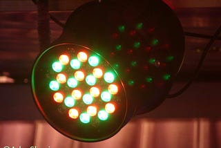 Close-up of a stage light with multiple LED bulbs emitting red and green light. The circular light fixture is mounted on a dark background, projecting colorful patterns onto a surface behind it.