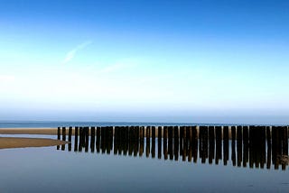 Landscape of Burgh-Haamstede, in the Netherlands, depicting a sandy beach with sea in the distance and shallow water in the foreground. There is a line of posts — wave breakers — driven into the sand.
