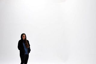 A girl with long brown hair and a blue shirt stands in a white room with a bright light at the top with her hands in a hoodie and a beanie cap on.