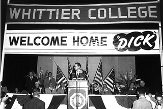 Black and white photo of Richard Nixon standing on stage in front of a podium with banners behind him reading “Whittier College” and “Welcome Home ‘Dick’.”