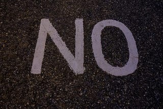Photograph of the word no spray-painted in white paint on black asphalt.